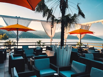 An outdoor terrace overlooking a serene lake surrounded by mountains. The patio is furnished with wicker chairs and sofas with blue cushions, shaded by large yellow and orange umbrellas. A palm tree is centrally placed, adding an exotic touch. White drapes hang around the edges, gently blowing in the breeze.