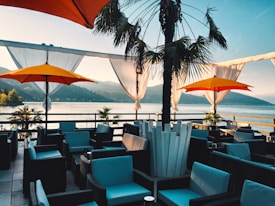 An outdoor terrace overlooking a serene lake surrounded by mountains. The patio is furnished with wicker chairs and sofas with blue cushions, shaded by large yellow and orange umbrellas. A palm tree is centrally placed, adding an exotic touch. White drapes hang around the edges, gently blowing in the breeze.