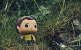 A Funko Pop figure with a yellow shirt and dark pants stands amidst tall, green grass. The background is a natural setting with blurred greenery, adding a sense of wilderness.