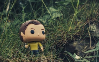 A Funko Pop figure with a yellow shirt and dark pants stands amidst tall, green grass. The background is a natural setting with blurred greenery, adding a sense of wilderness.