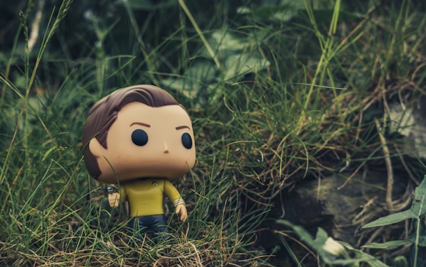 A Funko Pop figure with a yellow shirt and dark pants stands amidst tall, green grass. The background is a natural setting with blurred greenery, adding a sense of wilderness.