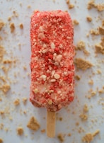 An ice cream bar covered in a layer of crumbly red and white coating is positioned on a flat surface. The bar has a wooden stick and is surrounded by scattered crumbs, possibly from the coating.