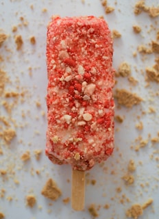An ice cream bar covered in a layer of crumbly red and white coating is positioned on a flat surface. The bar has a wooden stick and is surrounded by scattered crumbs, possibly from the coating.