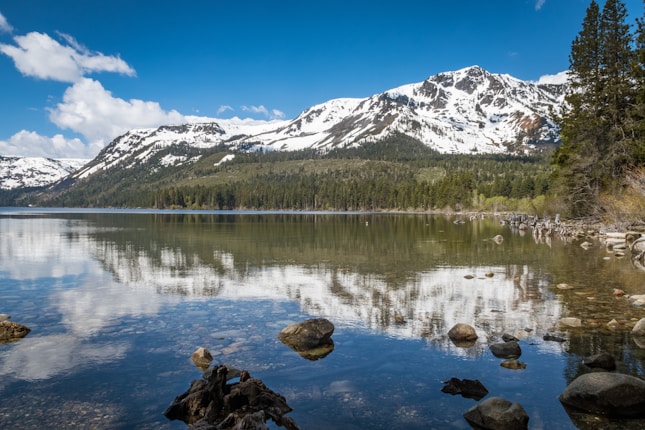 Snow-capped mountains are reflected in a calm, crystal-clear lake surrounded by a lush forest. The shoreline is dotted with rocks, and the sky is a bright blue with scattered fluffy clouds.