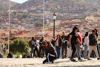 A bustling outdoor scene with a group of people gathered in a town square. In the foreground, a woman kneels while taking a photograph. Behind her, several individuals are walking and interacting, some are carrying backpacks and bottles of water. The background showcases a hillside densely packed with terracotta-roofed buildings, indicative of a hillside town. A lone street lamp stands out among the crowd.
