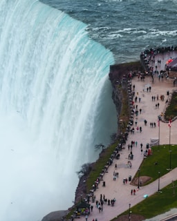 A breathtaking view of a massive waterfall cascading down with powerful force, surrounded by a vast body of water. A large number of people are gathered along the pathway on the right, observing the natural spectacle. The scene is bordered by lush green areas and features flags indicating a tourist destination.