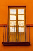 A wooden window with six glass panes is set into a bright, orange wall. Iron railings form a small balcony in front of the window, adding a decorative element.