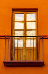 A wooden window with six glass panes is set into a bright, orange wall. Iron railings form a small balcony in front of the window, adding a decorative element.