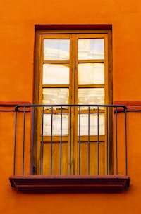 A wooden window with six glass panes is set into a bright, orange wall. Iron railings form a small balcony in front of the window, adding a decorative element.