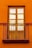 A wooden window with six glass panes is set into a bright, orange wall. Iron railings form a small balcony in front of the window, adding a decorative element.