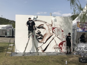A graffiti artist is actively working on a large mural featuring a stylized portrait using spray paint. The mural is set up outdoors with the artist using a ladder to reach higher parts of the artwork. The background includes festival tents and portable restrooms, suggesting a public or festival setting. The artist is wearing urban-style clothing, and there are spray paint cans on the ground.