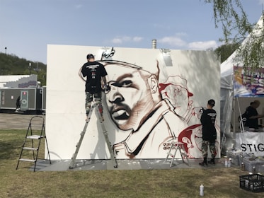 A graffiti artist is actively working on a large mural featuring a stylized portrait using spray paint. The mural is set up outdoors with the artist using a ladder to reach higher parts of the artwork. The background includes festival tents and portable restrooms, suggesting a public or festival setting. The artist is wearing urban-style clothing, and there are spray paint cans on the ground.