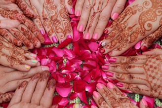 Multiple hands with intricate henna designs are arranged in a circular pattern over a bed of vibrant pink rose petals. The hands feature various traditional motifs, with most nails painted in shades of pink.