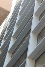 A modern building facade with a series of horizontal slats or louvers, creating a pattern of lines and shadows. The facade is composed of metallic and glass materials, reflecting light and capturing an urban architectural design.