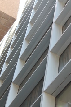 A modern building facade with a series of horizontal slats or louvers, creating a pattern of lines and shadows. The facade is composed of metallic and glass materials, reflecting light and capturing an urban architectural design.