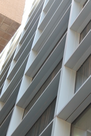 A modern building facade with a series of horizontal slats or louvers, creating a pattern of lines and shadows. The facade is composed of metallic and glass materials, reflecting light and capturing an urban architectural design.