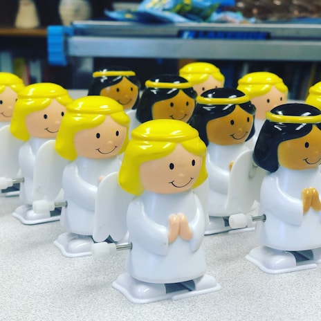 A collection of small plastic angel figurines with winding mechanisms, each featuring smiling faces, white robes, and wings. The angels have either blond or black hair and are arranged in rows on a light-colored surface, with some background elements slightly blurred.