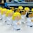 A collection of small plastic angel figurines with winding mechanisms, each featuring smiling faces, white robes, and wings. The angels have either blond or black hair and are arranged in rows on a light-colored surface, with some background elements slightly blurred.