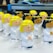 A collection of small plastic angel figurines with winding mechanisms, each featuring smiling faces, white robes, and wings. The angels have either blond or black hair and are arranged in rows on a light-colored surface, with some background elements slightly blurred.