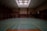 An empty indoor gymnasium with a green sports court featuring basketball hoops and multicolored lines for sporting activities. Overhead light fixtures illuminate the space through a patterned ceiling. The room is spacious, with muted tones and a somewhat subdued atmosphere.