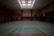 An empty indoor gymnasium with a green sports court featuring basketball hoops and multicolored lines for sporting activities. Overhead light fixtures illuminate the space through a patterned ceiling. The room is spacious, with muted tones and a somewhat subdued atmosphere.