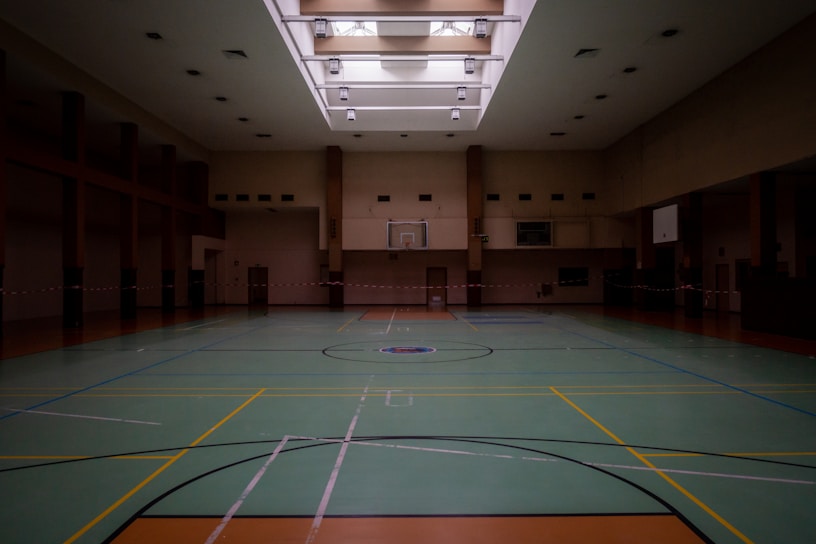 An empty indoor gymnasium with a green sports court featuring basketball hoops and multicolored lines for sporting activities. Overhead light fixtures illuminate the space through a patterned ceiling. The room is spacious, with muted tones and a somewhat subdued atmosphere.