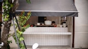 A small outdoor kitchen is visible, equipped with various cooking utensils and dishes. There is a tiled countertop with stacked bowls and a large cooking pot. A single person is seen working behind the counter. The space is partially covered by a dark awning, and leafy green branches from a tree are in the foreground.