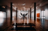A spacious art gallery with wooden floors features a large sculpture in the center, resembling a classical figure, and a silhouetted person standing near it. The gallery is well lit with overhead lights, and concrete pillars support the ceiling. Various artworks hang on the walls, including an abstract painting with a bright orange square.