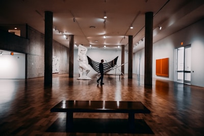 A spacious art gallery with wooden floors features a large sculpture in the center, resembling a classical figure, and a silhouetted person standing near it. The gallery is well lit with overhead lights, and concrete pillars support the ceiling. Various artworks hang on the walls, including an abstract painting with a bright orange square.