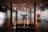 A spacious art gallery with wooden floors features a large sculpture in the center, resembling a classical figure, and a silhouetted person standing near it. The gallery is well lit with overhead lights, and concrete pillars support the ceiling. Various artworks hang on the walls, including an abstract painting with a bright orange square.