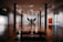 A spacious art gallery with wooden floors features a large sculpture in the center, resembling a classical figure, and a silhouetted person standing near it. The gallery is well lit with overhead lights, and concrete pillars support the ceiling. Various artworks hang on the walls, including an abstract painting with a bright orange square.