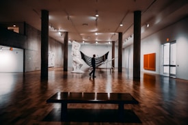 A spacious art gallery with wooden floors features a large sculpture in the center, resembling a classical figure, and a silhouetted person standing near it. The gallery is well lit with overhead lights, and concrete pillars support the ceiling. Various artworks hang on the walls, including an abstract painting with a bright orange square.