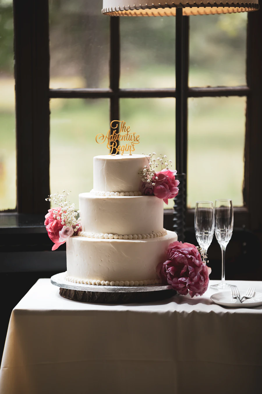 Bolo de três andares branco com flores cor-de-rosa ao lado de duas taças de espumante