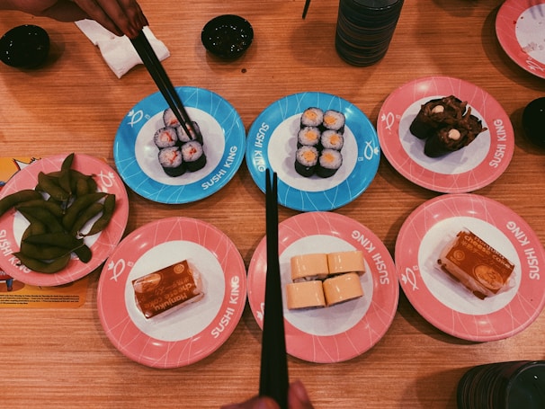 A variety of sushi dishes are arranged on colorful plates on a wooden table. Several plates have sushi rolls, including ones wrapped in seaweed and others with rice on the outside. There are also plates with edamame and dessert items. Chopsticks are held above one of the dishes, ready to pick up sushi.