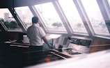 A person wearing a uniform stands in a ship's control room, looking out large, slanted windows. The control panel is visible, with buttons, switches, and a microphone. Outside, a blurry cityscape can be seen through the raindrop-covered windows, adding an element of distance and contemplation.