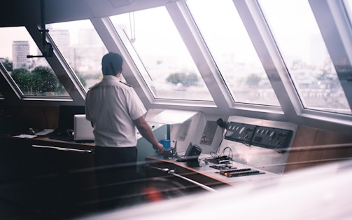 A person wearing a uniform stands in a ship's control room, looking out large, slanted windows. The control panel is visible, with buttons, switches, and a microphone. Outside, a blurry cityscape can be seen through the raindrop-covered windows, adding an element of distance and contemplation.