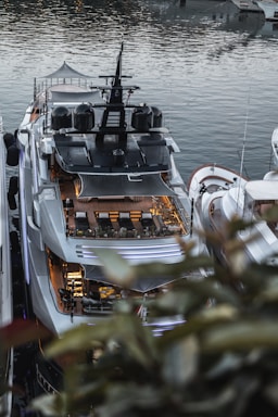 An overhead view of a large luxury yacht docked in a marina. The yacht features multiple decks, with luxurious seating arrangements and modern design elements. Other boats are visible on either side, and the water reflects the surrounding vessels.