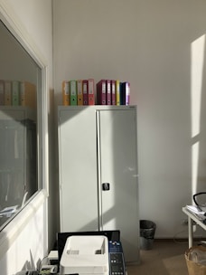 A well-lit office corner features a tall gray cabinet topped with colorful binders. Sunlight streams in from the side, casting shadows on the wall. In the foreground, a printer sits next to a desk filled with office supplies. A trash bin is seen near the wall.