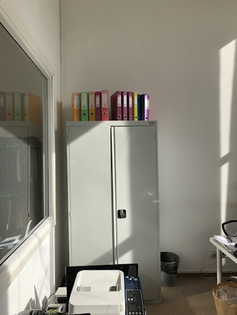 A well-lit office corner features a tall gray cabinet topped with colorful binders. Sunlight streams in from the side, casting shadows on the wall. In the foreground, a printer sits next to a desk filled with office supplies. A trash bin is seen near the wall.