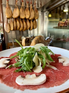 A close-up of a dish featuring thinly sliced raw meat, likely beef carpaccio, garnished with fresh arugula, shaved parmesan cheese, and mushroom slices. In the background, a row of cured hams hang from the ceiling, suggesting a deli or charcuterie setting.