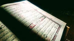 An open book displaying Arabic calligraphy on its pages. The background is dark, and the book appears to be illuminated by a light source coming from the upper left corner. The pages include decorative elements with red and black ink, and there are verses or text blocks within ornate borders.