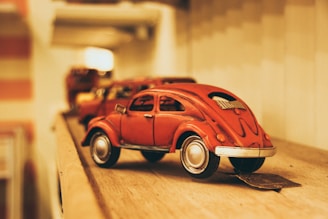A vintage red toy car is placed on a wooden surface, surrounded by a warm, muted background with soft lighting. The focus is on the detailed design of the car, which has an antique feel and nostalgic aesthetic.