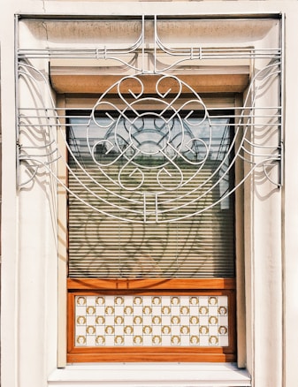 A window adorned with an intricate metal grille featuring curved and circular patterns. The metalwork is symmetrical and elegant, with a floral-like design at the center. The window has horizontal blinds partially open, allowing a view of a wooden frame and a decorative panel with checkered tiles below. The tiles exhibit a repeated ornate motif.