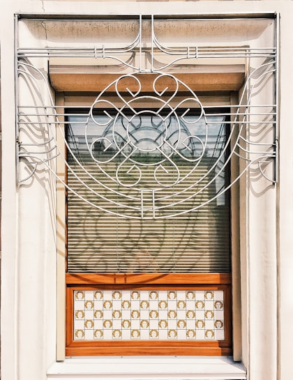 A window adorned with an intricate metal grille featuring curved and circular patterns. The metalwork is symmetrical and elegant, with a floral-like design at the center. The window has horizontal blinds partially open, allowing a view of a wooden frame and a decorative panel with checkered tiles below. The tiles exhibit a repeated ornate motif.