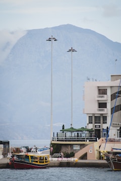 A waterfront scene featuring a building with balconies and a rooftop terrace. Two tall lampposts stand in front of the building. A small, colorful boat is docked by the pier, with a larger mountain visible in the background under a cloudy sky.
