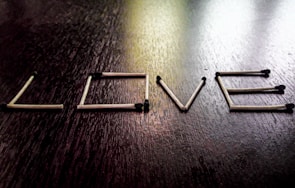 A series of matchsticks arranged to form the word 'LOVE' on a dark wooden surface. Each matchstick is carefully positioned to create the illusion of letters. The matches have blackened tips, suggesting they are unlit.