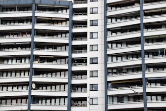A modern apartment building with multiple floors, each featuring balconies with railings. The building has a repetitive architectural style with a mix of glass windows and white panels separated by vertical gray columns.