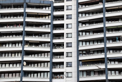A modern apartment building with multiple floors, each featuring balconies with railings. The building has a repetitive architectural style with a mix of glass windows and white panels separated by vertical gray columns.