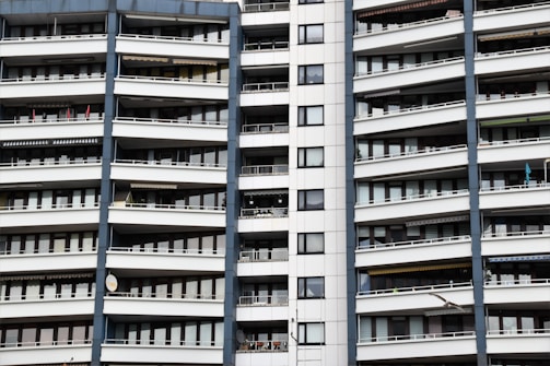 A modern apartment building with multiple floors, each featuring balconies with railings. The building has a repetitive architectural style with a mix of glass windows and white panels separated by vertical gray columns.