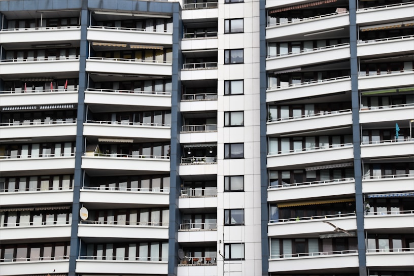 A modern apartment building with multiple floors, each featuring balconies with railings. The building has a repetitive architectural style with a mix of glass windows and white panels separated by vertical gray columns.